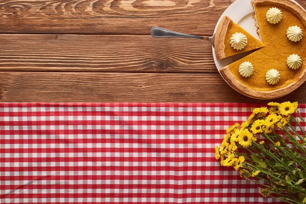 Torta di zucca tagliata con panna montata vicino a mazzo di fiori gialli e tovaglia a quadri su tavolo di legno — Foto stock