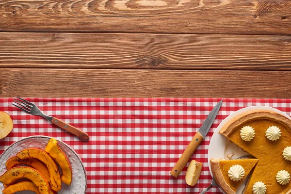 Torta di zucca tagliata vicino al piatto con zucca al forno affettata, forchetta e coltello sulla tovaglia a quadri sul tavolo di legno — Foto stock