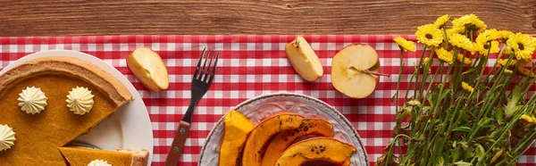 Colpo panoramico di torta di zucca vicino alla zucca affettata al forno, fiori gialli, mela tagliata e forchetta su tovaglia a quadri su superficie di legno — Foto stock