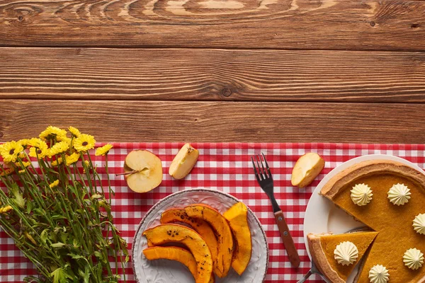 Pumpkin pie with whipped cream near sliced baked pumpkin, yellow flowers, cut apple and fork on plaid tablecloth on wooden surface — Stock Photo