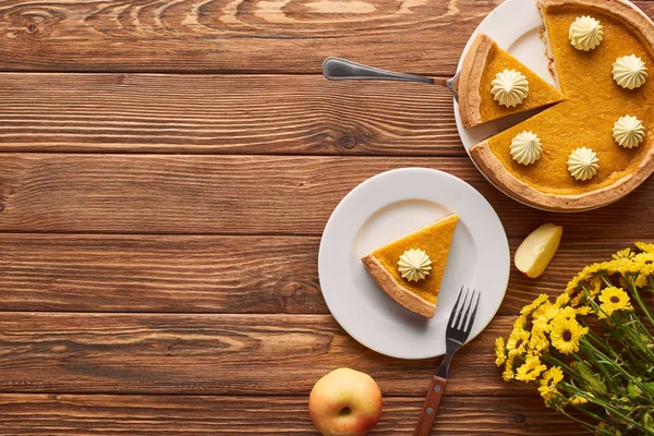Cut pumpkin pie with whipped cream, apple, and yellow flowers on wooden surface — Stock Photo