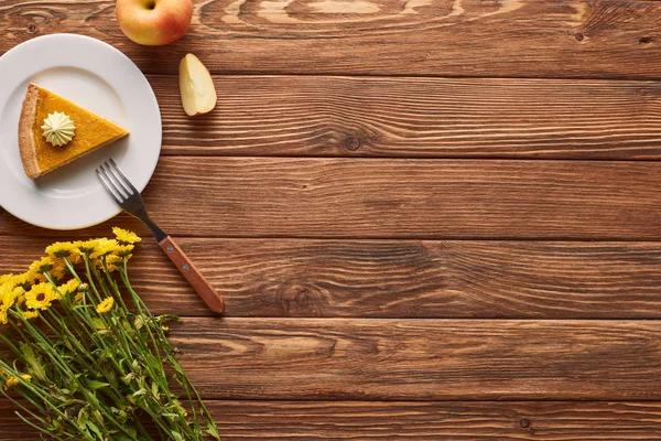 Piece of pumpkin pie with whipped cream near fork, apple, and yellow flowers on wooden surface — Stock Photo