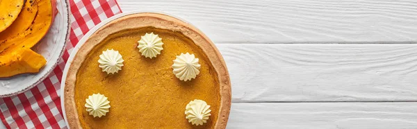 Colpo panoramico di torta di zucca con panna montata sulla tovaglia a quadretti vicino alla zucca al forno tagliata a fette sulla superficie di legno bianca — Foto stock