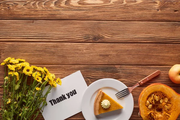 Morceau de tarte à la citrouille avec crème fouettée près de carte de souhaits de remerciement, fourchette, citrouille crue et fleurs jaunes sur table en bois — Photo de stock
