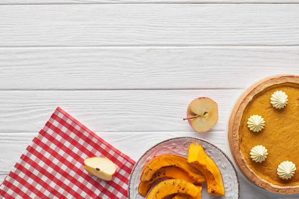 Leckere Kürbiskuchen mit Schlagsahne in der Nähe gebackener Kürbisscheiben, geschnittener Apfel und karierte Serviette auf weißer Holzoberfläche — Stockfoto