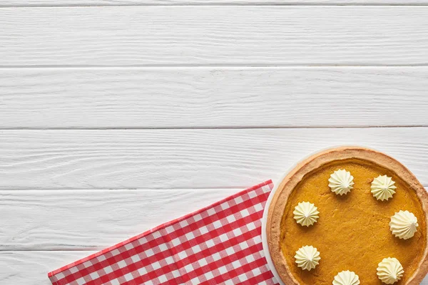 Délicieuse tarte à la citrouille avec crème fouettée près de serviette à carreaux sur surface en bois blanc — Photo de stock