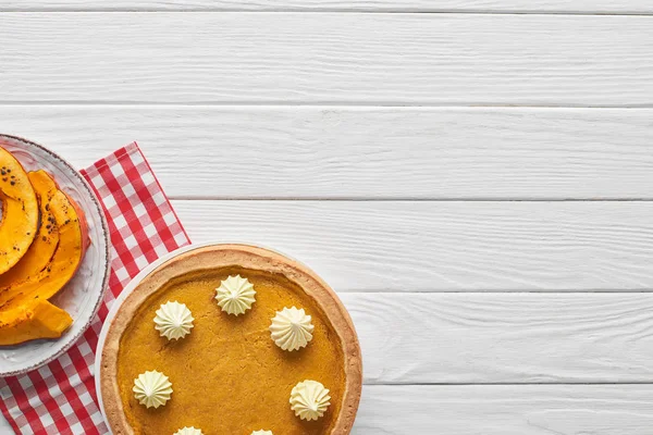 Tasty pumpkin pie with whipped cream on checkered napkin near baked sliced pumpkin on white wooden surface — Stock Photo
