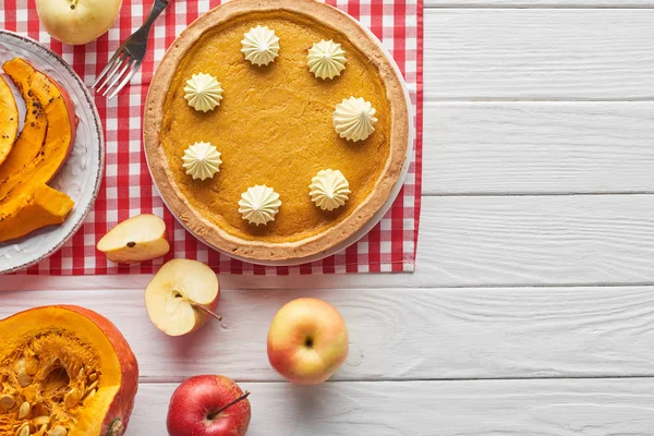 Délicieuse tarte à la citrouille avec crème fouettée sur une serviette à carreaux près de citrouilles crues et cuites au four, pommes coupées et entières, et fourchette sur une surface en bois blanc — Photo de stock