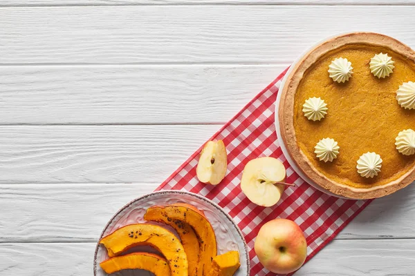 Вкусный тыквенный пирог со взбитыми сливками на клетчатой салфетке рядом с запечённой тыквой, вырезать и целые яблоки на белом деревянном столе — стоковое фото