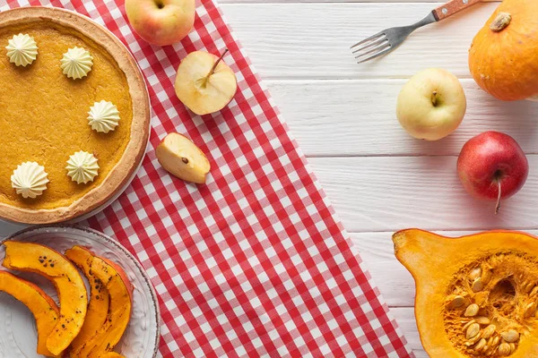 Delicioso pastel de calabaza con crema batida en servilleta a cuadros cerca de calabazas horneadas y crudas, manzanas enteras y cortadas, y tenedor en mesa de madera blanca - foto de stock