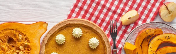 Panoramic shot of pumpkin pie with whipped cream on plaid napkin near baked and raw pumpkins, cut apple and fork on white woode table — Stock Photo