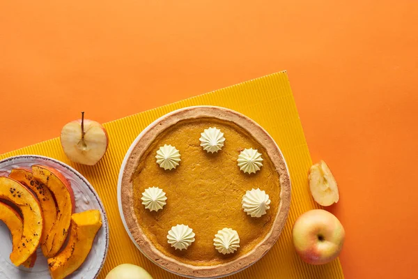 Вкусный тыквенный пирог со взбитыми сливками рядом с запечённой тыквой, целые и разрезанные яблоки на оранжевой поверхности — стоковое фото