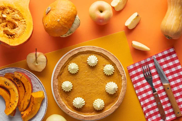 Leckere Kürbiskuchen mit Schlagsahne in der Nähe von gebackenen und rohen Kürbissen, ganzen und geschnittenen Äpfeln, Gabel und Messer auf orangefarbener Oberfläche — Stockfoto
