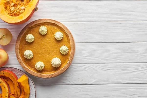 Tasty pumpkin pie with whipped cream near baked and raw pumpkins, whole and cut apples on white wooden table — Stock Photo
