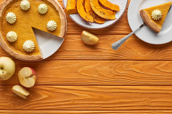 Leckere Kürbiskuchen mit Schlagsahne neben gebackenem Kürbis, ganze und geschnittene Äpfel auf orangefarbenem Holztisch — Stockfoto