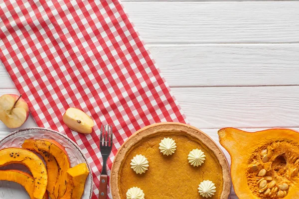 Tasty pumpkin pie with whipped cream on checkered napkin near baked and raw pumpkins, cut apple and fork on white wooden table — Stock Photo