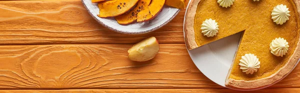 Plan panoramique de délicieuse tarte à la citrouille avec crème fouettée près de la citrouille cuite au four et pomme coupée sur une table en bois orange — Photo de stock