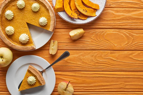 Delicioso pastel de calabaza con crema batida cerca de calabaza al horno, manzanas enteras y cortadas en una mesa de madera naranja - foto de stock