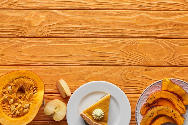 Morceau de tarte à la citrouille savoureuse avec crème fouettée près de citrouilles cuites au four et crues, et couper la pomme sur une table en bois orange — Photo de stock