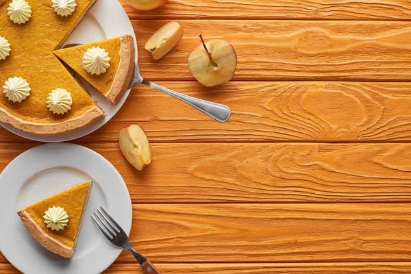 Pastel de calabaza sabroso y cortado con crema batida en el plato con espátula, tenedor y manzana cortada en una mesa de madera marrón - foto de stock