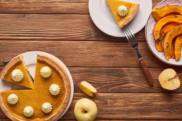 Pastel de calabaza cortado con crema batida cerca de calabaza al horno, manzanas enteras y cortadas y tenedor en mesa de madera marrón - foto de stock