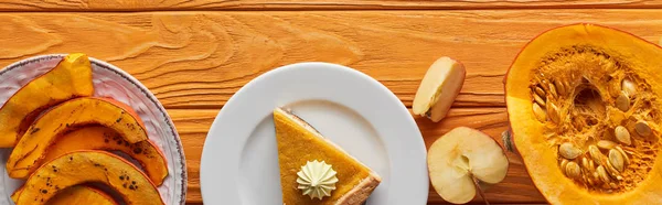 Панорамний знімок смачного гарбузового пирога зі збитими вершками біля запечених і сирих гарбузів, і нарізати яблуко на апельсиновому дерев'яному столі — стокове фото