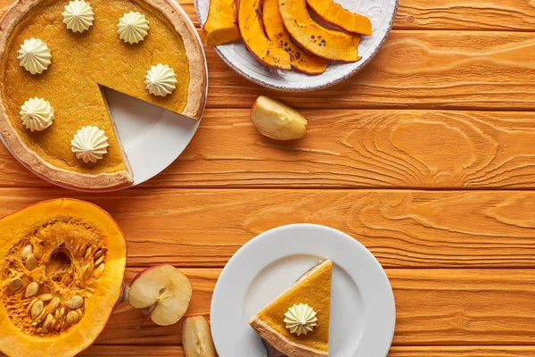 Вкусный, нарезанный тыквенный пирог со взбитыми сливками рядом с запечёнными и сырыми тыквами, и яблоко на оранжевом деревянном столе — стоковое фото