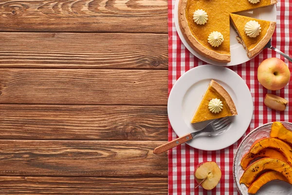 Delicioso pastel de calabaza con crema batida en mantel a cuadros cerca de calabaza al horno, tenedor, corte y manzanas enteras en mesa de madera marrón — Stock Photo