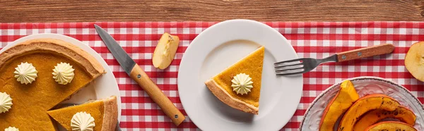 Panoramic shot of pumpkin pie with whipped cream on checkered tablecloth near baked pumpkin, fork, knife, and cut apples on brown wooden table — Stock Photo