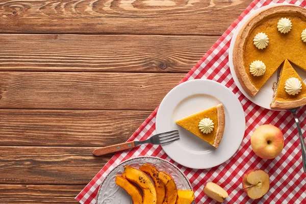 Tasty pumpkin pie with whipped cream on checkered tablecloth near baked pumpkin, cut and whole apples on brown wooden table — Stock Photo