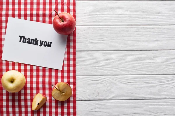 Vue du dessus des pommes mûres et carte de remerciement sur table blanche en bois avec serviette à carreaux rouge — Photo de stock