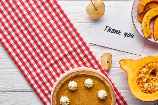 Vista dall'alto di torta di zucca, mele mature e biglietto di ringraziamento su tavolo bianco in legno con tovagliolo a quadri — Foto stock