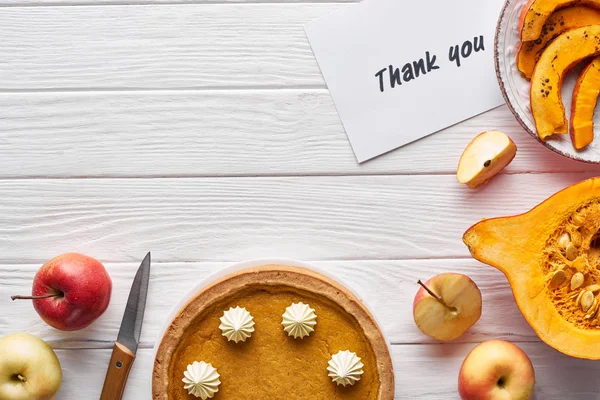 Vue du dessus de tarte à la citrouille, pommes mûres et carte de remerciement sur table blanche en bois — Photo de stock