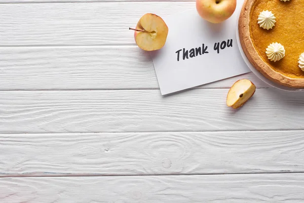 Vue du dessus de tarte à la citrouille, pommes mûres et carte de remerciement sur table blanche en bois — Photo de stock