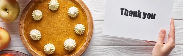 Vista recortada de la mujer sosteniendo tarjeta de agradecimiento cerca de pastel de calabaza en la mesa blanca de madera con manzanas, tiro panorámico - foto de stock