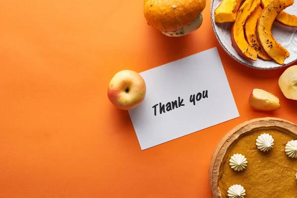 Vista superior de pastel de calabaza, manzanas maduras y gracias tarjeta sobre fondo naranja - foto de stock