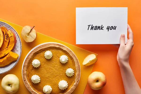 Cropped view of woman holding thank you card near pumpkin pie, ripe apples on orange background — Stock Photo