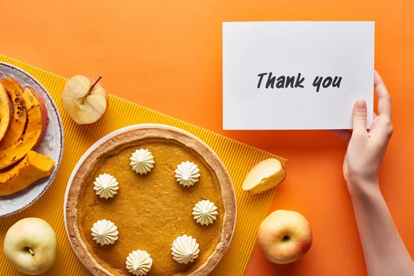 Vista recortada de la mujer sosteniendo gracias tarjeta cerca de pastel de calabaza, manzanas maduras sobre fondo naranja - foto de stock