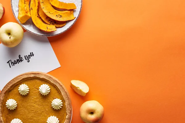 Vista superior de pastel de calabaza, manzanas maduras y gracias tarjeta en fondo naranja con espacio para copiar - foto de stock