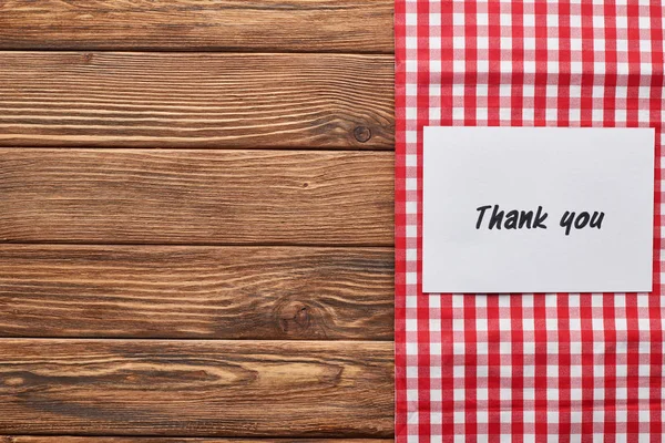 Vista superior de la tarjeta de agradecimiento en la mesa marrón de madera con servilleta a cuadros rojo - foto de stock