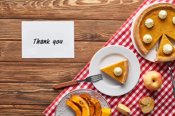 Vista dall'alto di torta di zucca, mele mature e biglietto di ringraziamento su tavolo marrone in legno con tovagliolo a quadri rosso — Foto stock