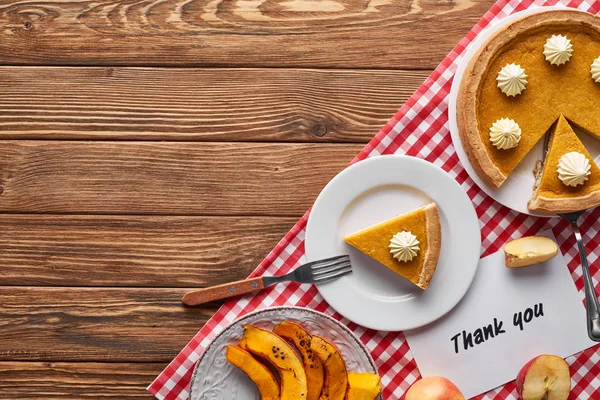 Vista superior del pastel de calabaza, manzanas maduras y tarjeta de agradecimiento en la mesa marrón de madera con servilleta a cuadros roja - foto de stock