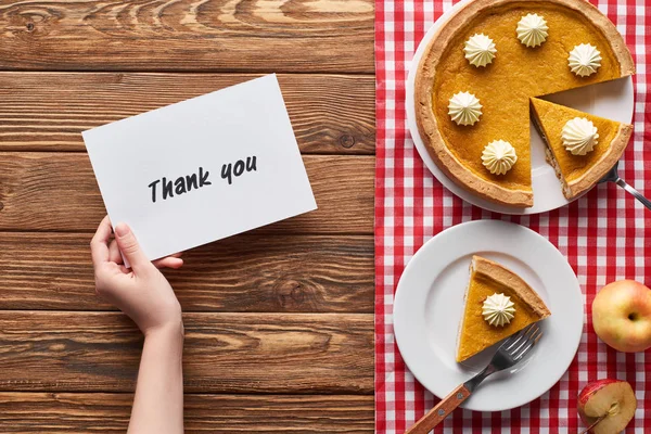 Cropped view of woman holding thank you card near pumpkin pie, ripe apples on plaid napkin on wooden table — Stock Photo