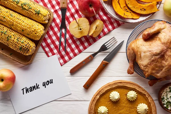 Vue du dessus de tarte à la citrouille, dinde et légumes servis à la table blanche en bois avec carte de remerciement pour le dîner de Thanksgiving — Photo de stock