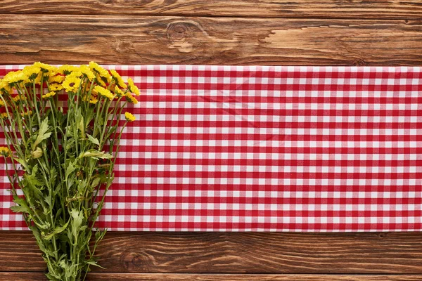 Top view of yellow wildflowers on red checkered napkin at wooden table — Stock Photo