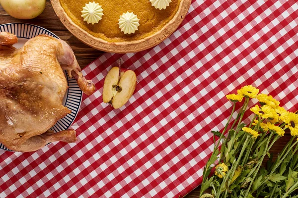 Top view of pumpkin pie and roasted turkey on red checkered napkin with yellow flowers at wooden table for Thanksgiving dinner — Stock Photo