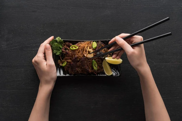 Vista recortada de la mujer comiendo deliciosos fideos tailandeses picantes con palillos en la superficie gris de madera - foto de stock