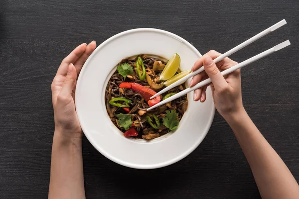 Cropped view of woman holding chopsticks near plate with tasty spicy thai noodles on wooden grey surface — Stock Photo