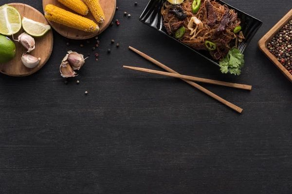 Вид зверху смачної гострої тайської локшини біля паличок та свіжих інгредієнтів на дерев'яній сірій поверхні з місцем для тексту — стокове фото