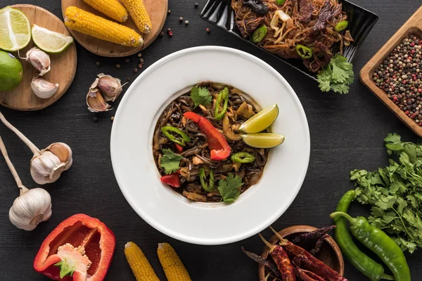 Vista dall'alto di gustose tagliatelle di carne piccante tailandese vicino a bacchette e ingredienti freschi sulla superficie grigia di legno — Foto stock